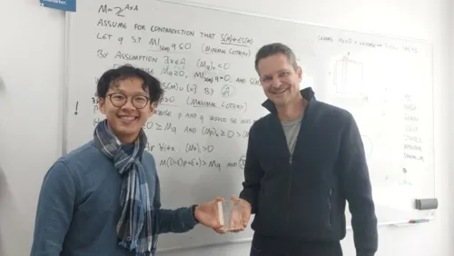 Chris Dong reicht dem Preisträger, Prof. Brandt, die Trophäe. Im Hintergrund ist ein vollgeschriebenes Whiteboard zu sehen.